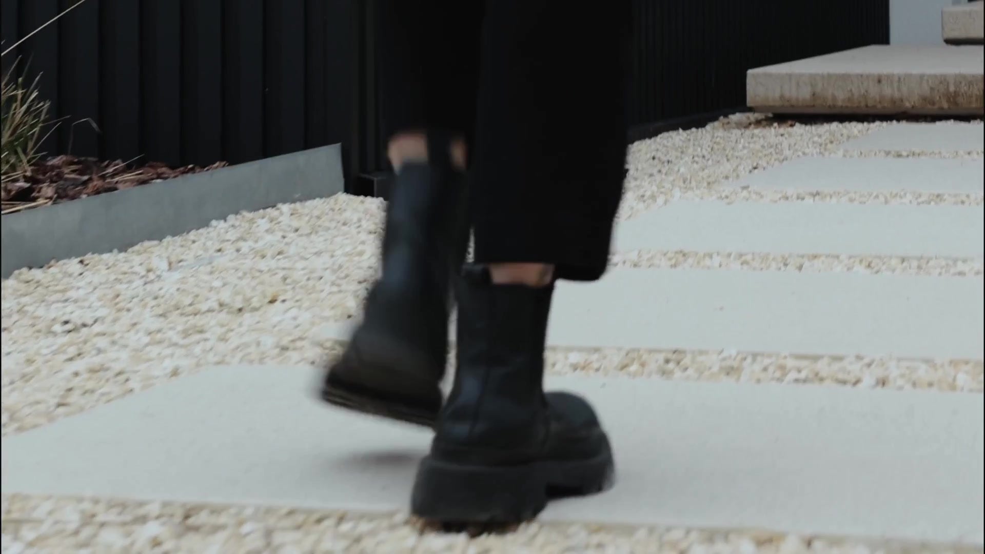Person in black boots walking on a gravel and stone path along a black slatted garage wall